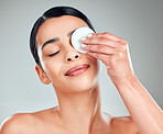 A beautiful happy mixed race woman using a cotton pad to remove makeup during a grooming routine. Hispanic woman applying cleanser to her face against grey copyspace background in a studio