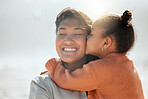 Adorable mixed race girl kisses her mother on the cheek and outs her arms around her while at the beach. Little daughter showing mommy love and affection while she closes her her eyes and smile