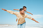 Cheerful young mixed race father being playful while giving his son a piggyback ride on his back and stretching out their arms pretending to fly. Young man and his boy having fun and spending time together while on vacation by the beach