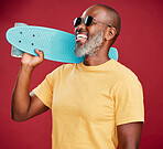 One mature african american man standing with a mini skateboard in studio isolated against a red background. Handsome and carefree man wearing sunglasses and laughing happily. He is ready for summer