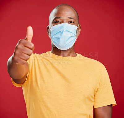 Buy stock photo Black man, portrait and mask with thumbs up for winning, protection or good safety on a red studio background. African, male person or model with face cover, like emoji or yes sign for great review