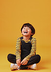 A cute little asian boy sitting on the floor with casual clothes while laughing and crossing his legs against an orange copyspace  background. Adorable happy little boy safe and alone