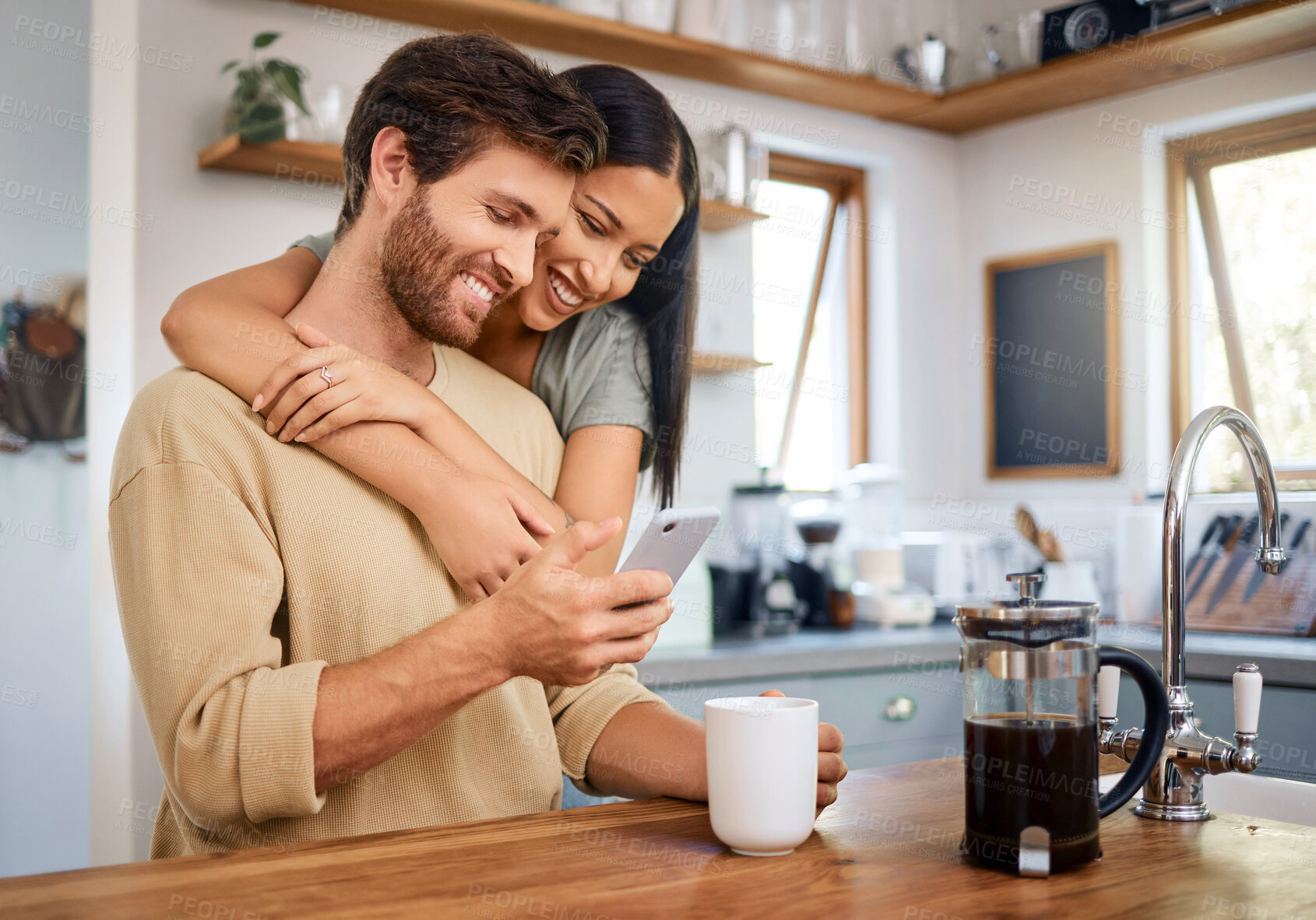 Buy stock photo Happy young interracial couple being loving and affectionate at home. Young man using his smartphone and holds coffee cup while his girlfriend embraces him from behind. Browsing social media or sending text message