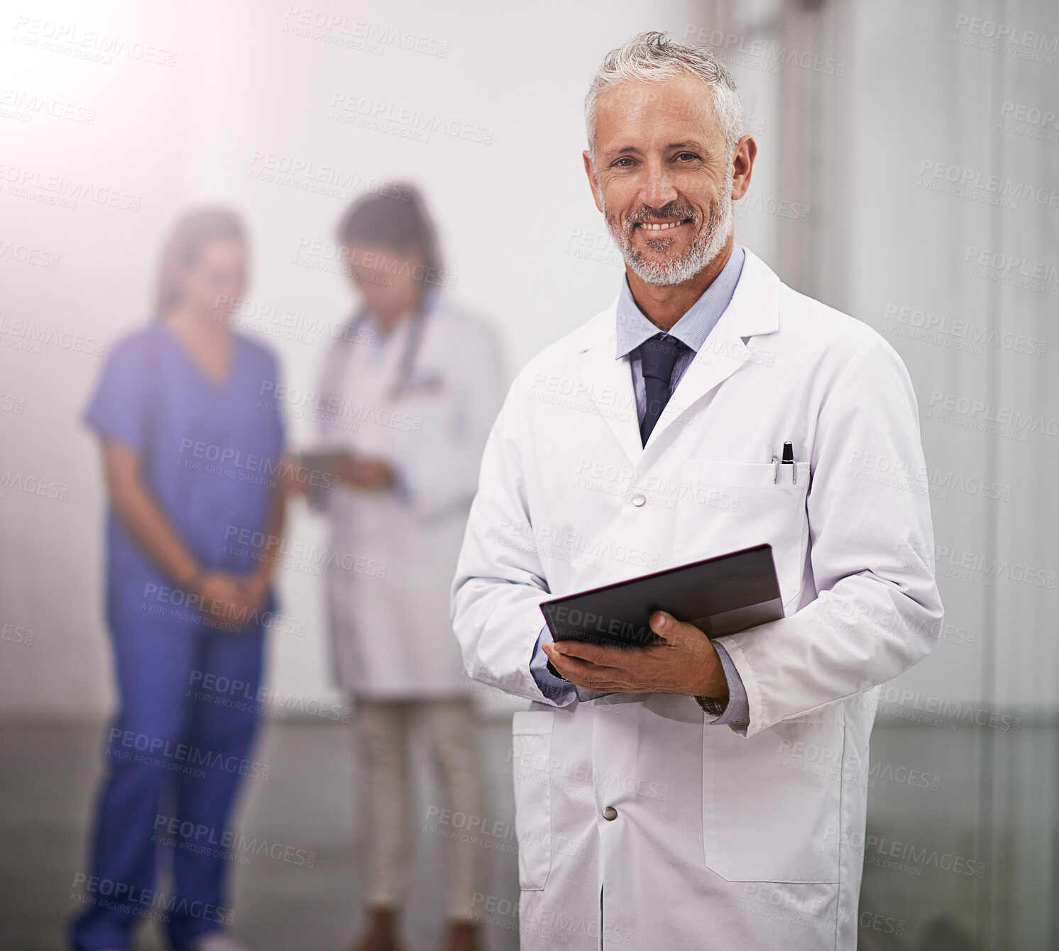 Buy stock photo Cropped portrait of a mature doctor standing in the hospital corridor with two colleagues in the background