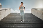 Young mixed race female athlete listening to music on headphones while running down the steps of a building outside. Young female focused on her speed, body, fitness and cardio health while training