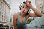 Portrait of a young mixed race female athlete wearing wireless headphones and listening to music while standing outside in the city wiping her sweaty forehead and tired from a workout. Determined to get a fit and toned body