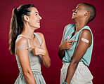 Covid vaccinated African american and mixed race women showing, pointing at arm plaster. Two happy people isolated on red studio background with copyspace. Black woman and hispanic with corona vaccine