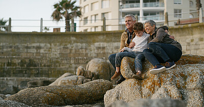 Buy stock photo Grandparents, child and happy on rocks at beach with hug, memory and outdoor on holiday with bonding. Senior man, woman and boy with embrace, care and love with family on vacation with view in Italy