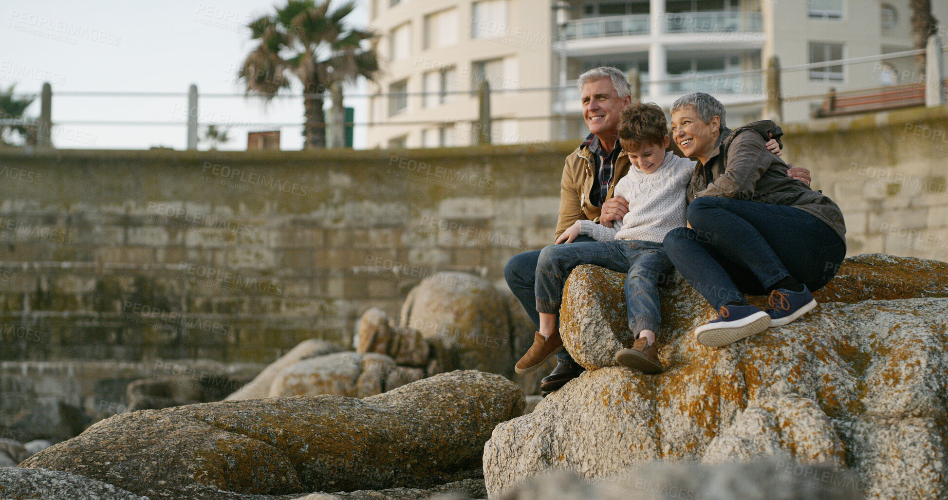 Buy stock photo Grandparents, child and happy on rocks at beach with hug, memory and outdoor on holiday with bonding. Senior man, woman and boy with embrace, care and love with family on vacation with view in Italy