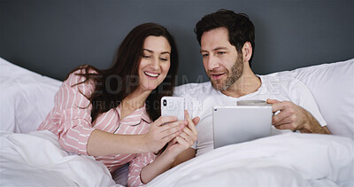 Buy stock photo Cellphone, tablet and couple relaxing in bed in home for networking, connectivity or browsing online. Happy, rest and young people with phone and digital technology in bedroom for contact in house.