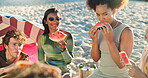 People, friends and eating with watermelon at beach, picnic and conversation with reunion on vacation. Group, women and men with fruit, happy and outdoor together with diversity by ocean in sunshine