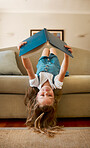 Carefree content caucasian little girl reading a book alone sitting upside down on the couch at home. One cheerful positive child holding and reading a story