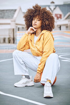 Buy stock photo Trendy, stylish and fashionable female basketball player sitting on a ball at the court against an urban background. Young, edgy and serious woman with an afro posing outside in the city in the day