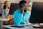 One young african american call centre telemarketing agent talking on a headset while working on a computer in an office. Focused businessman consultant operating a helpdesk for customer service support