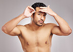 Handsome young mixed race man posing shirtless in studio isolated against a grey background. Hispanic male plucking his eyebrows with a tweezers. All part of his daily grooming and skincare routine