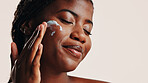 Happy, black woman and lotion on face in studio for facial hydration, treatment and sunscreen protection. Female person, apply cream and spf cosmetics for dermatology, anti aging and white background