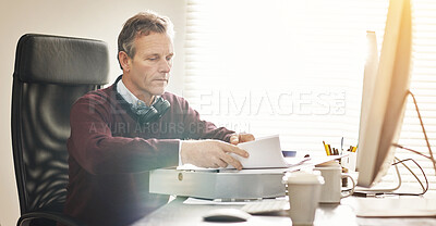 Buy stock photo A businessman analysing paperwork and using a computer in a modern office. A mature male executive working online and going over a business document at his desk