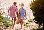 Couple, walking and beach with picnic for date, outdoor vacation or summer holiday on sandy shore. Back view of man and woman enjoying stroll with basket and coolerbox for sunny day by ocean coast