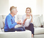 Mature couple drinking tea and eating sandwiches while having a conversation and relaxing together on a sofa at home