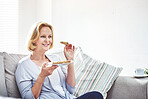 Happy mature woman eating a sandwich and drinking tea while relaxing on a sofa at home