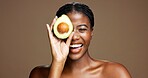 Skincare, happy and black woman with avocado in studio with natural, antioxidants or face glow. Smile, beauty and African female person with fruit for moisturizing dermatology by brown background.
