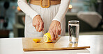 Hands, cutting lemon and glass of water in kitchen for nutrition, vitamin c or detox for wellness. Person, citrus fruit and lemonade for hydration, diet or benefits for health with drink at apartment