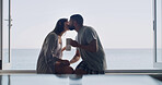 Kiss, love and coffee with couple at window of home for connection, romance and peace. Relax, bonding and commitment with man and woman and drink tea in apartment for marriage, space and morning