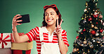 Christmas, peace sign and woman with selfie in studio for festive, tradition or holiday celebration. Happy, phone and person with photography picture by xmas tree and gifts by green background.
