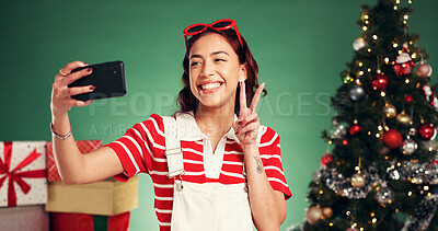 Buy stock photo Christmas, peace sign and woman with selfie in studio for festive, tradition or holiday celebration. Happy, phone and person with photography picture by xmas tree and gifts on green background.