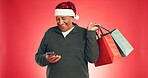 Phone, shopping bags and senior man with christmas in studio with networking on mobile app. Laugh, gifts and elderly male person with cellphone for communication with xmas season by red background.