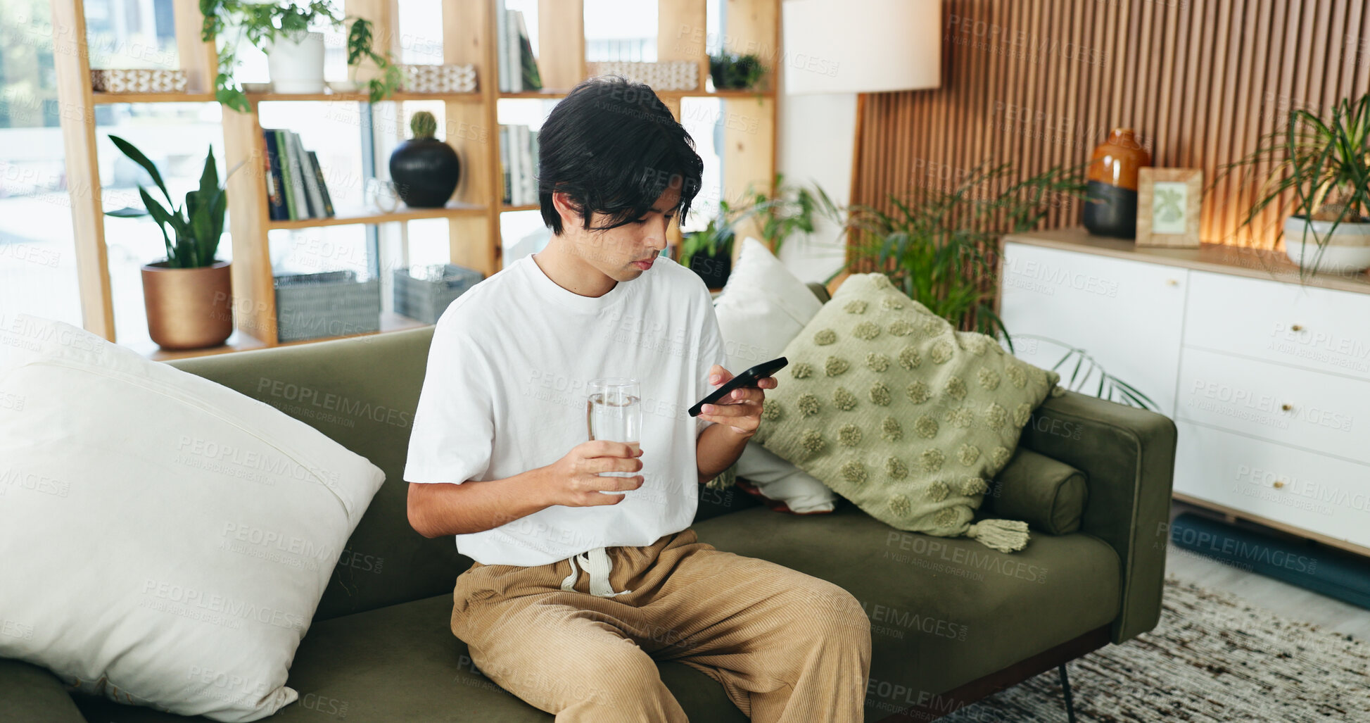 Buy stock photo Phone, relax and water with Asian man on sofa in home living room for hydration or social media. Break, drink and glass with person scrolling mobile app for browsing, connection or online search