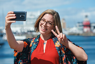 Buy stock photo Woman, selfie and happy at waterfront with peace sign on vacation, travel and glasses on web by ocean. Person, smile and photography with app, social media and profile picture on seaside holiday