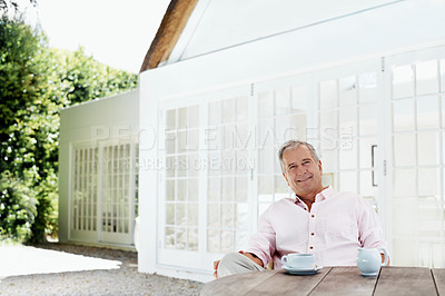 Buy stock photo A senior man smiling at the camera while sitting outside his house with a cup of coffee in front of him
