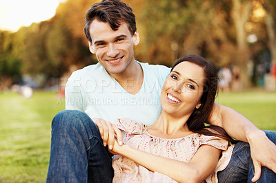 Buy stock photo Lovely young couple smiling at you while relaxing in a park on a sunny evening