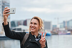 Beautiful travel woman taking selfie photo using smartphone camera in waterfront harbour making peace sight with hand sharing vacation holding mobile phone