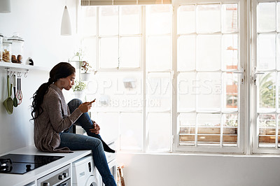 Buy stock photo Shot of a young woman using her cellphone in the kitchen