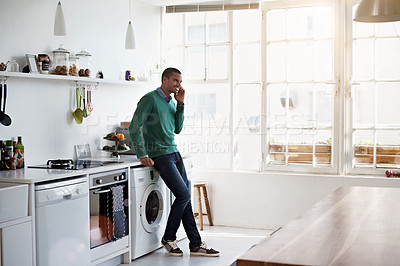 Buy stock photo Shot of a young man leaning on his kitchen counter talking on the phone