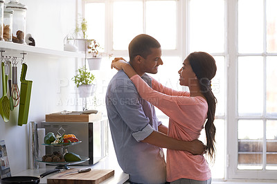 Buy stock photo Shot of a young couple talking while standing arm in arm together in their kitchen