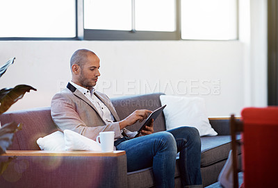 Buy stock photo Shot of a young man sitting on a sofa in an office using a digital tablet