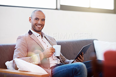 Buy stock photo Portrait of a young man sitting on a sofa in an office drinking coffee and using a digital tablet