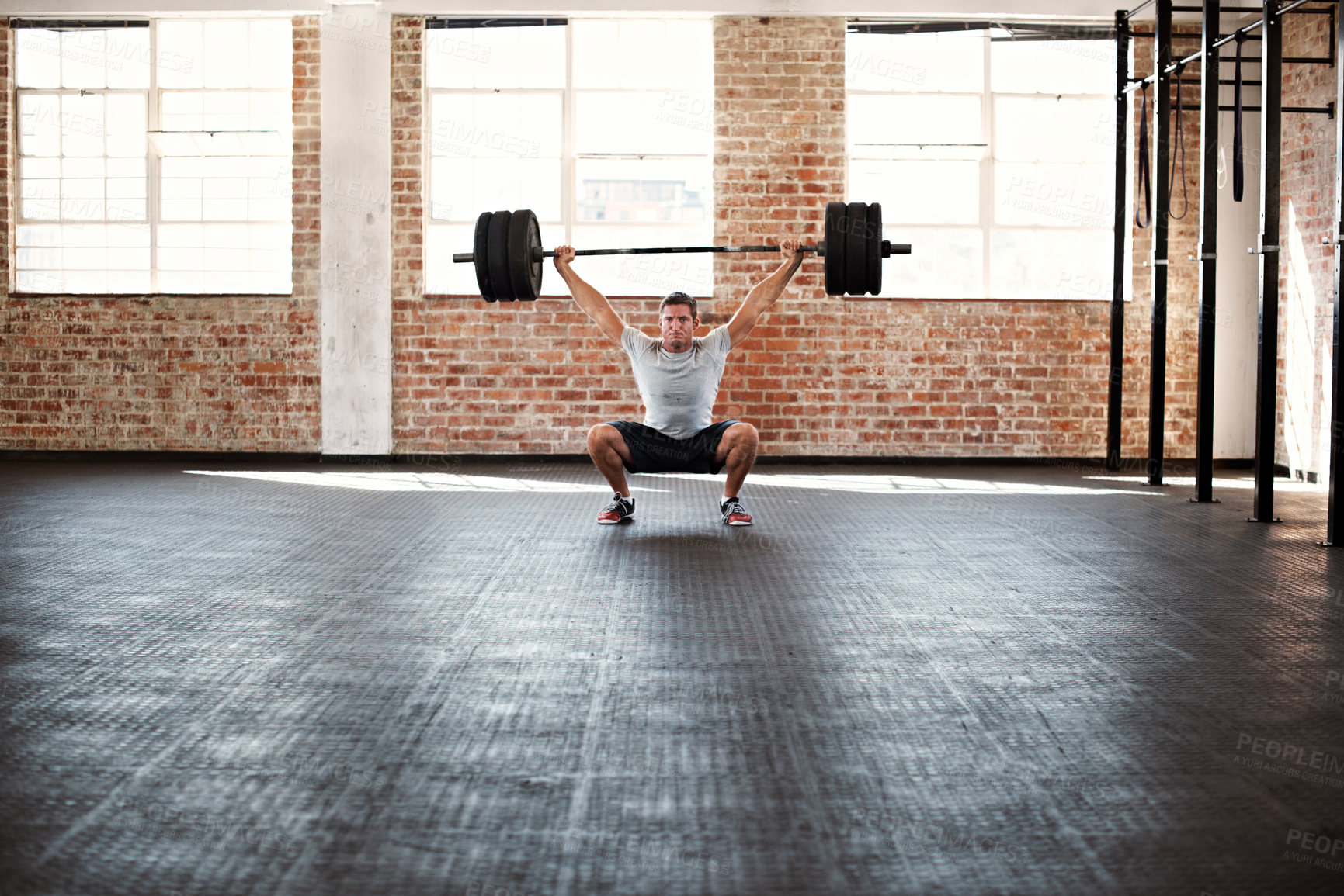 Buy stock photo Portrait of a muscular young man in a gym doing squats while holding a barbell