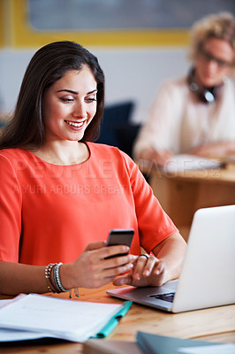 Buy stock photo Shot of a designer texting at her desk with her colleague blurred in the background