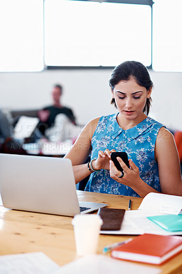 Buy stock photo Shot of a designer texting at her desk with her colleague blurred in the background