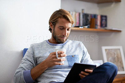 Buy stock photo Shot of a young man using a digital tablet and drinking a coffee while relaxing at home