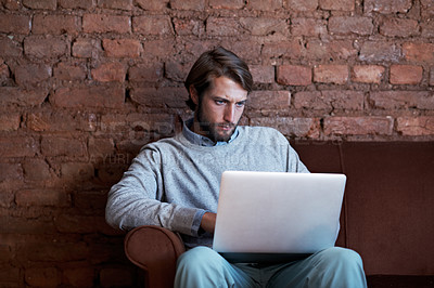 Buy stock photo Shot of a young man working on a laptop while sitting on a sofa at home