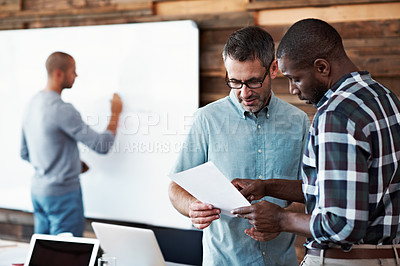 Buy stock photo Cropped shot of two businessmen talking while a colleague prepares a presentation in the background