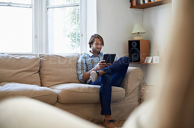 Buy stock photo Shot of a young man using a digital tablet while sitting on the sofa at home