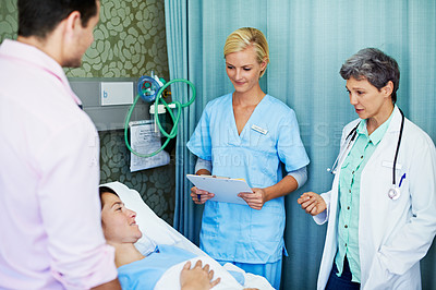 Buy stock photo Shot of doctors talking to a woman lying in a hospital bed while her husband looks on