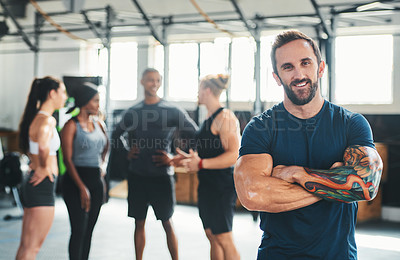 Buy stock photo Cropped portrait of a handsome young male athlete standing with his arms folded in the gym with other gym-goers in the background