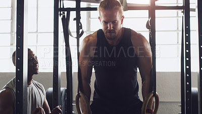 Buy stock photo Shot of a young man exercising with gymnastic rings with the help of a trainer in a gym